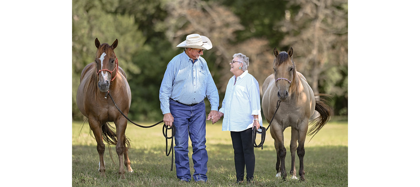 An older couple stands hand-in-hand in a sunny pasture, each holding the lead of a horse.